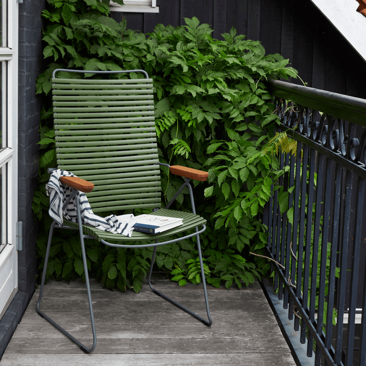 A green outdoor lounge chair with bamboo armrest set on a wooden deck balcony with metal railing and lush green plants