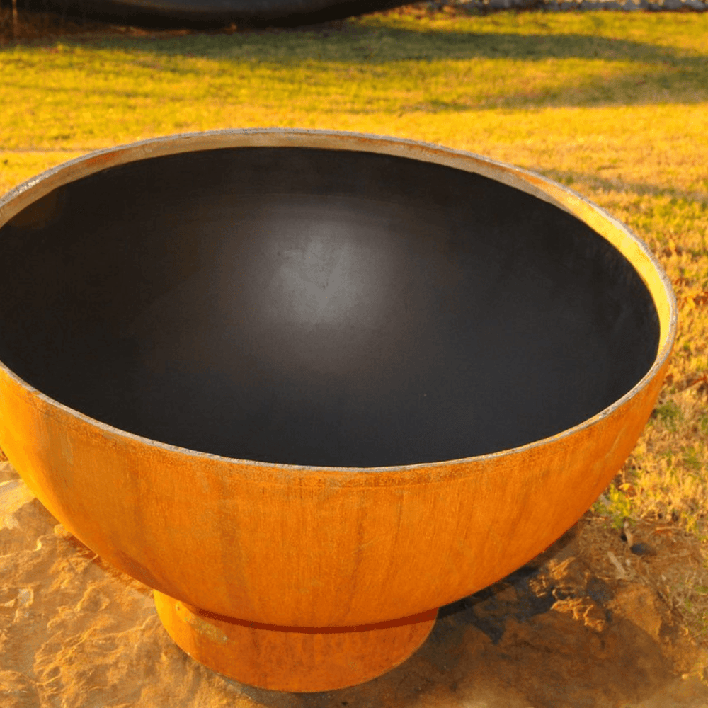 Metal fire pit bowl in orange rust color with placed on concrete surface beside grassy field