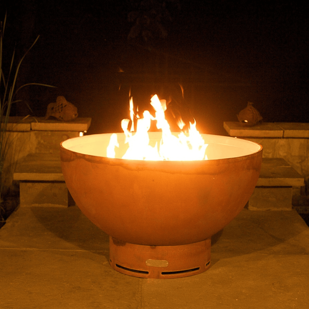 Metal fire pit bowl in orange rust color with bright flame placed on an outdoor patio during nighttime