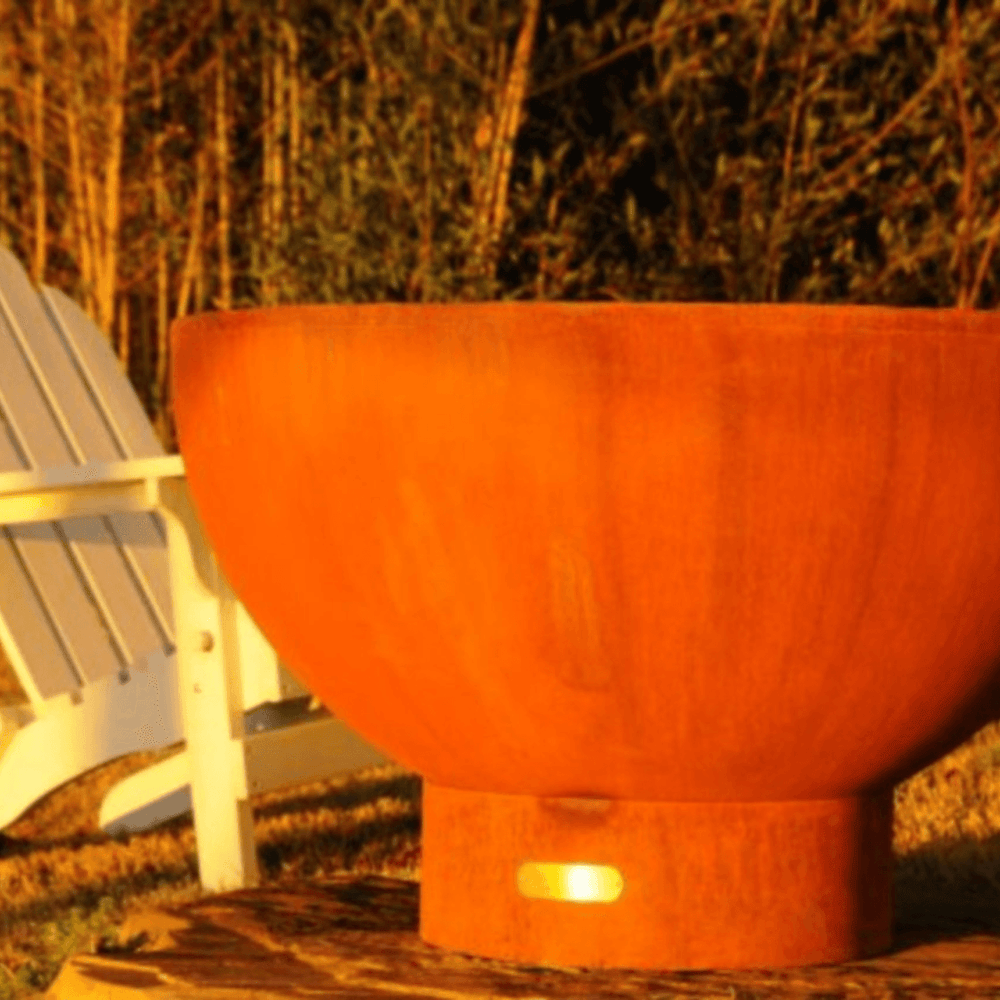 Metal fire pit bowl in orange rust color with placed on concrete surface beside white wooden chair surrounded by grass and trees